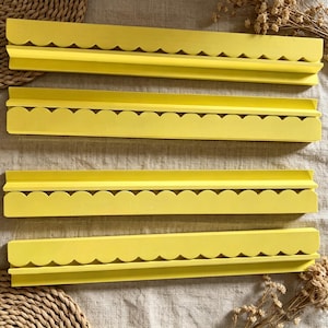 May include: Four long, bright yellow wall shelves, each featuring a decorative scalloped edge along the front. These horizontal display ledges have a flat top surface and a raised back lip, designed for mounting to a wall to hold small items.