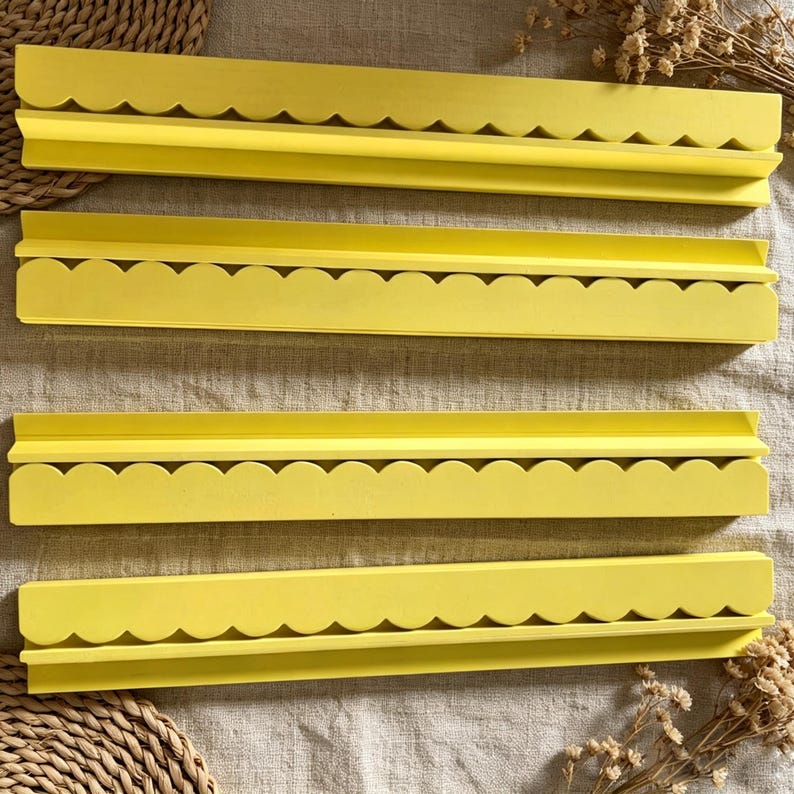 May include: Four long, bright yellow wall shelves, each featuring a decorative scalloped edge along the front. These horizontal display ledges have a flat top surface and a raised back lip, designed for mounting to a wall to hold small items.