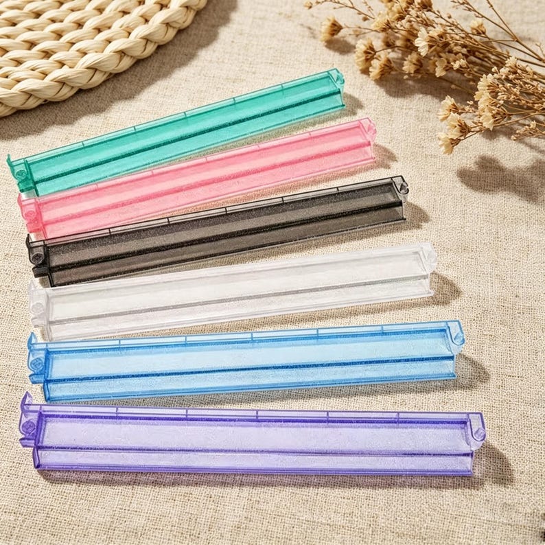 May include: A set of six rectangular plastic bag sealing clips in translucent green, pink, black, clear, blue, and purple. Each clip features a hinged design with a long, narrow channel for securing bags, providing a functional tool for kitchen storage.