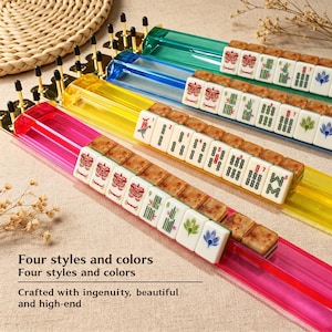 May include: Four translucent Mahjong tile racks in pink, yellow, blue, and green, each featuring gold-toned metal end caps with vertical pins. The racks hold white Mahjong tiles with various green, red, and blue engraved symbols and patterns.