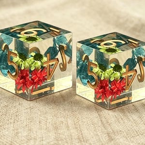 May include: Two clear resin dice featuring embedded dried flowers in shades of red, lime green, and teal. Each die has gold-colored numbers etched into the surface, including a prominent five and four on the visible faces, with a smooth finish.
