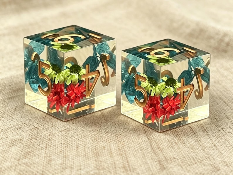 May include: Two clear resin dice featuring embedded dried flowers in shades of red, lime green, and teal. Each die has gold-colored numbers etched into the surface, including a prominent five and four on the visible faces, with a smooth finish.