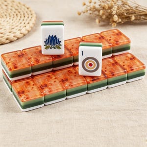May include: A collection of rectangular mahjong tiles featuring a layered design with a white base, a green middle section, and a translucent amber-colored top. Two tiles stand upright, one displaying a blue lotus flower and the other a target symbol.