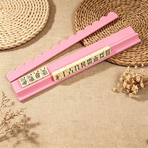 May include: A pink Mahjong tile rack with a scalloped top edge, holding a row of ten off-white Mahjong tiles. Each tile features distinct green, red, and black character and symbol engravings. The rack is designed with a horizontal groove to hold the tiles upright during gameplay.