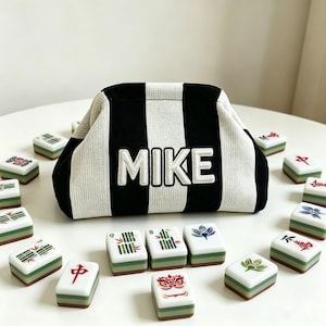 May include: A black and white striped knit pouch with a frame closure, featuring the name MIKE embroidered in white block letters on the front, surrounded by a collection of rectangular mahjong tiles with various green and red symbols.