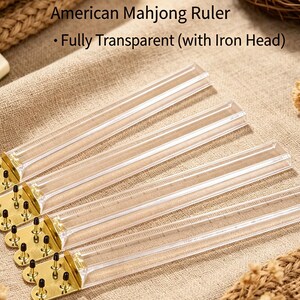 May include: Four American Mahjong rulers made of clear, transparent plastic. Each ruler features a gold-toned metal end piece with four small, black-tipped metal pins. The text "American Mahjong Ruler - Fully Transparent (with Iron Head)" is visible.