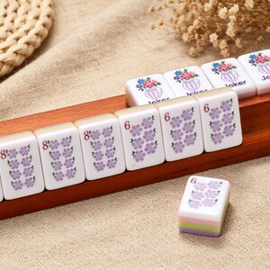 May include: A set of white Mahjong tiles with purple floral patterns and numerical markings, along with four tiles featuring a colorful flower vase graphic and the word Joker, all arranged on a long wooden rack with a grooved surface.