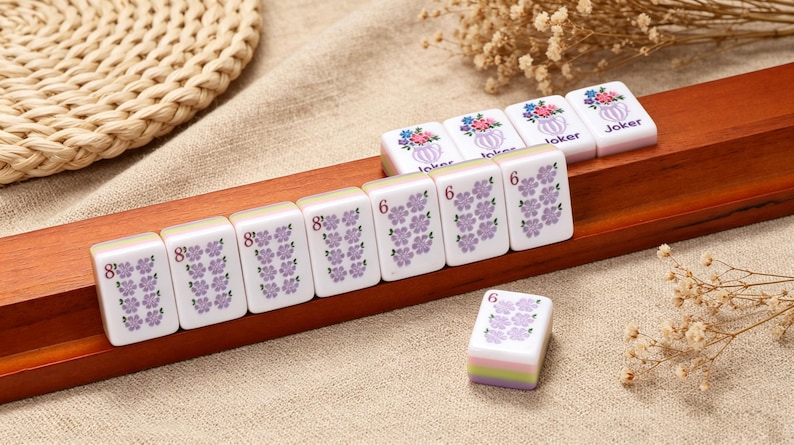 May include: A set of white Mahjong tiles with purple floral patterns and numerical markings, along with four tiles featuring a colorful flower vase graphic and the word Joker, all arranged on a long wooden rack with a grooved surface.