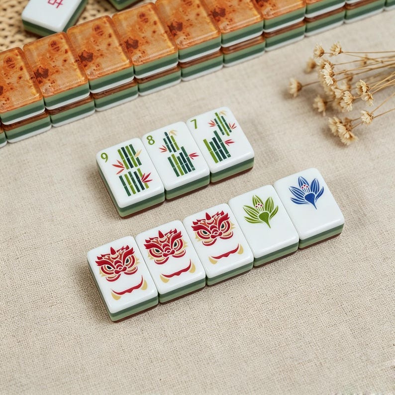May include: A set of rectangular mahjong tiles featuring white faces with green bamboo designs numbered 7, 8, and 9, alongside tiles displaying red lion masks, a green floral motif, and a blue lotus flower, with stacked tiles in the background.