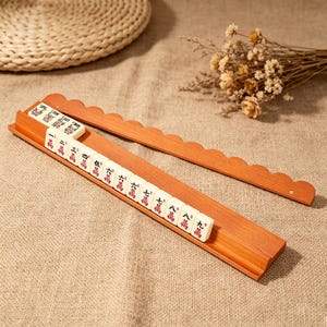 May include: A wooden mahjong tile rack with a scalloped top edge, holding a row of white tiles with red and black Chinese characters. The rack is made of light brown wood and features a grooved base to keep the tiles upright during gameplay.