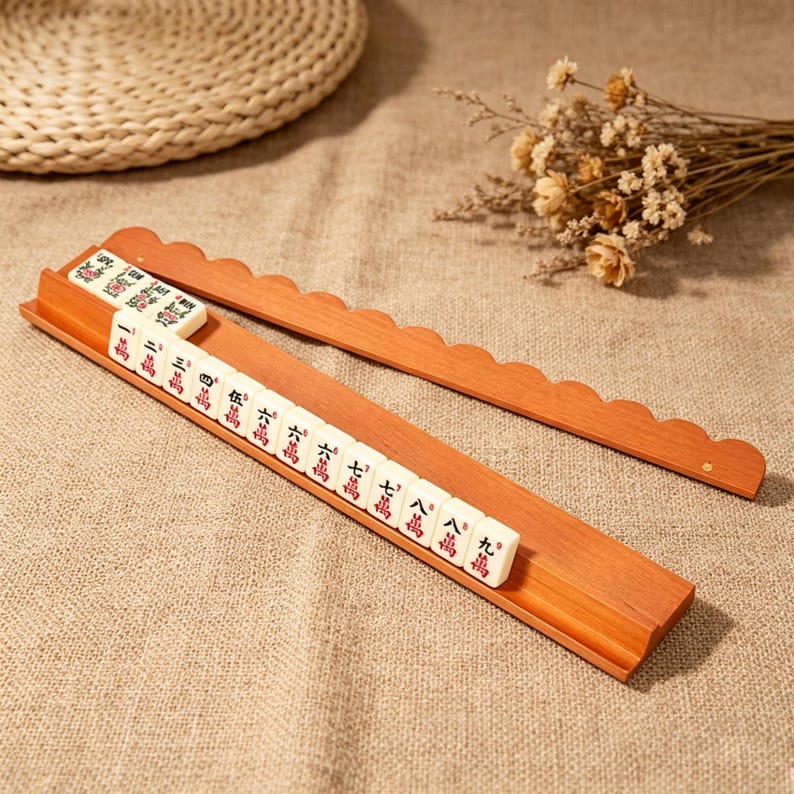 May include: A wooden mahjong tile rack with a scalloped top edge, holding a row of white tiles with red and black Chinese characters. The rack is made of light brown wood and features a grooved base to keep the tiles upright during gameplay.