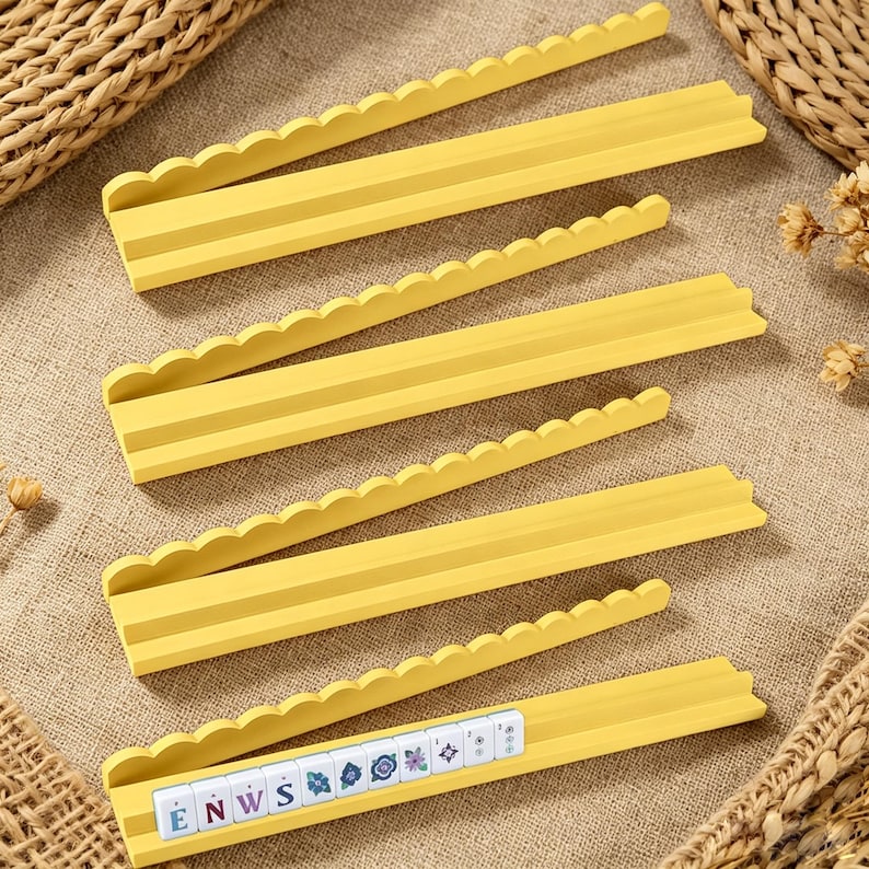 May include: Four yellow game tile racks with a scalloped top edge and a tiered design. The bottom rack holds a row of white square tiles featuring blue letters and floral patterns, spelling out the letters E, N, W, and S followed by decorative icons.