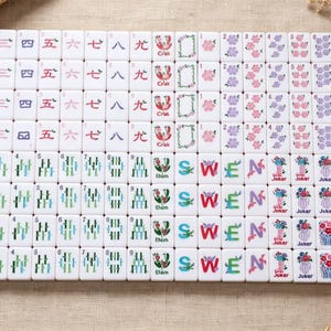 May include: A set of white rectangular mahjong tiles arranged in a grid. The tiles feature various colorful illustrations including Chinese numerals, bamboo stalks, birds, flowers, and the letters S, W, E, N, along with the text Crab, Joker, and D&acirc;m.
