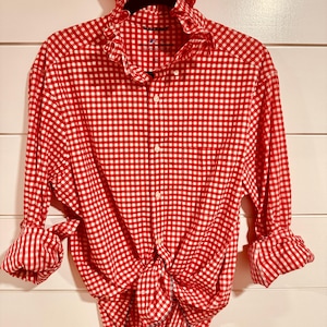 Puede incluir: Una camisa de manga larga con botones y un patrón de cuadros vichy en rojo y blanco. La camisa tiene un cuello con volantes, una tapeta de botones frontal y mangas remangadas. El dobladillo inferior está atado en un nudo en la parte delantera.