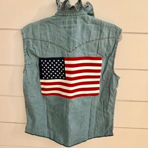 May include: A light blue sleeveless denim vest featuring a ruffled collar and a rectangular patch of the United States flag stitched onto the center of the back. The vest has a classic western-style yoke seam across the upper back area.