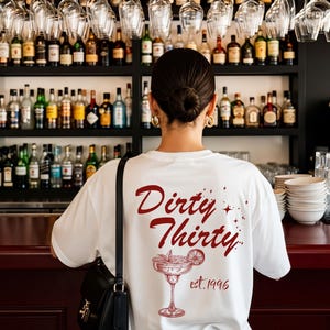 May include: A white t-shirt featuring a dark red graphic on the back. The design includes the text "Dirty Thirty" in a cursive font, small star accents, a line-art illustration of a margarita glass with a lime slice, and the text "est. 1996".