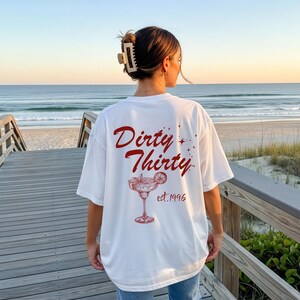 May include: A white short-sleeve t-shirt featuring a red graphic on the back. The text reads Dirty Thirty and est. 1996, accompanied by an illustration of a cocktail glass with a citrus slice garnish and small star accents scattered around the design.