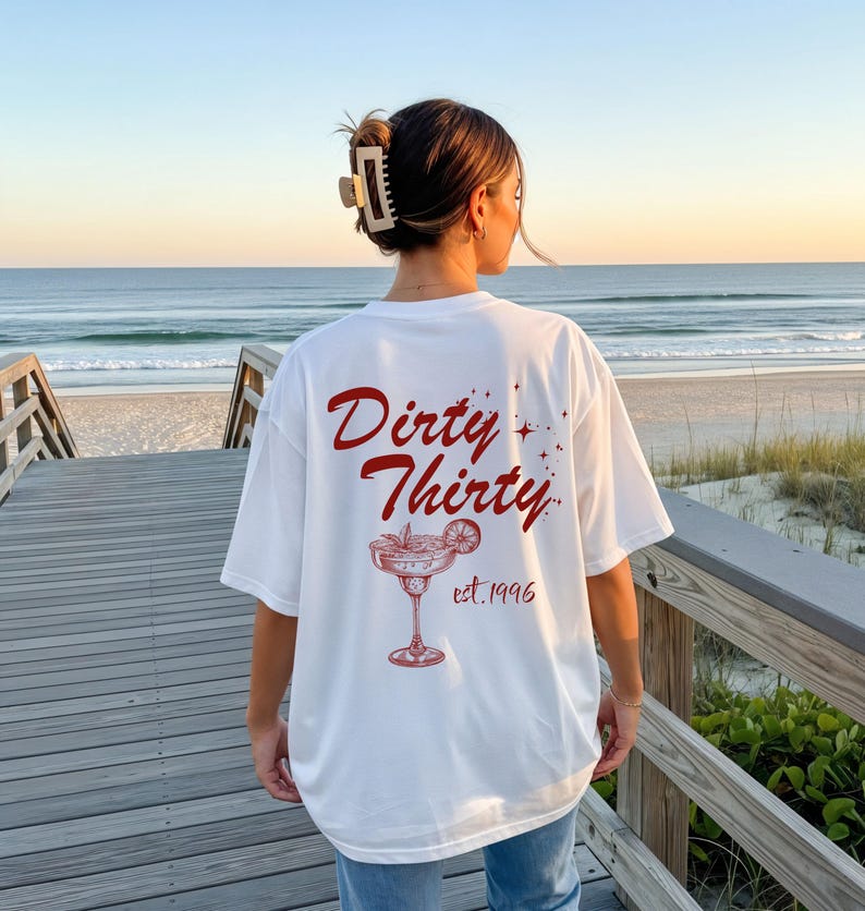 May include: A white short-sleeve t-shirt featuring a red graphic on the back. The text reads Dirty Thirty and est. 1996, accompanied by an illustration of a cocktail glass with a citrus slice garnish and small star accents scattered around the design.
