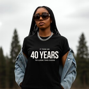 May include: Black t-shirt with the text "IT TOOK ME 40 YEARS TO LOOK THIS GOOD" in white. The person is wearing a denim jacket, sunglasses, and a silver chain necklace. The background is a blurred outdoor scene.