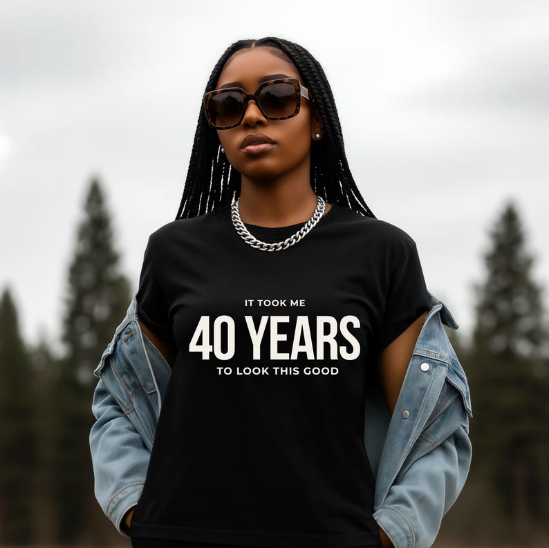 May include: Black t-shirt with the text "IT TOOK ME 40 YEARS TO LOOK THIS GOOD" in white. The person is wearing a denim jacket, sunglasses, and a silver chain necklace. The background is a blurred outdoor scene.