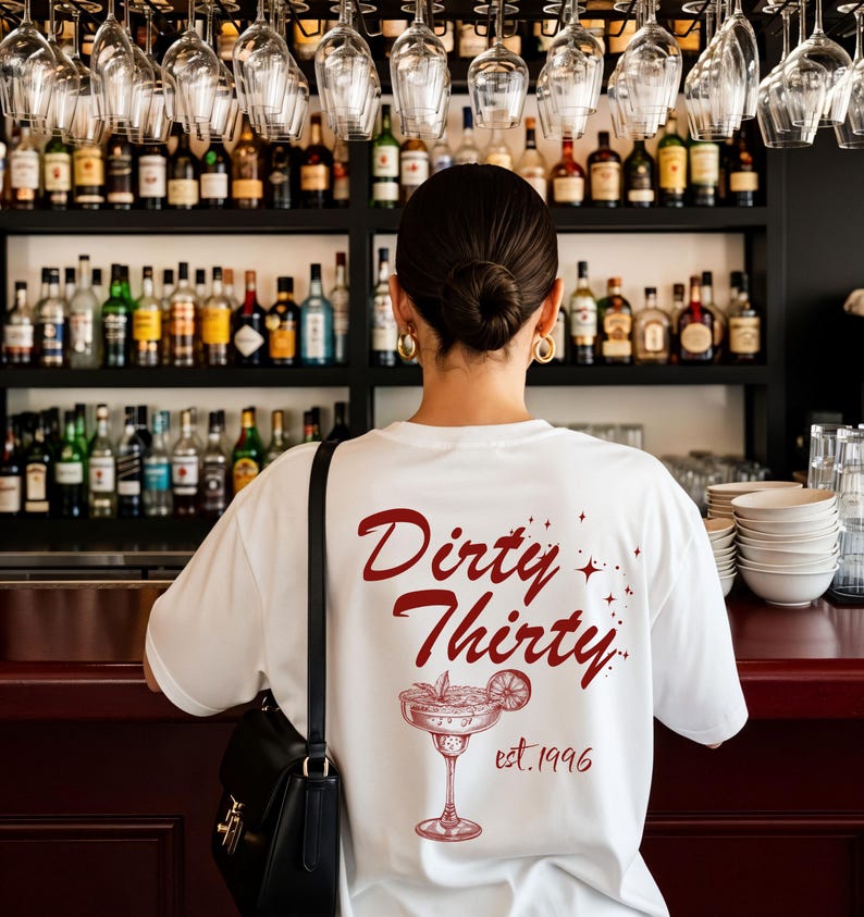 May include: A white t-shirt featuring a dark red graphic on the back. The design includes the text "Dirty Thirty" in a cursive font, small star accents, a line-art illustration of a margarita glass with a lime slice, and the text "est. 1996".