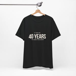 May include: Black t-shirt with the text "IT TOOK ME 40 YEARS TO LOOK THIS GOOD" in white. The shirt is hanging on a wooden hanger attached to a metal bar.
