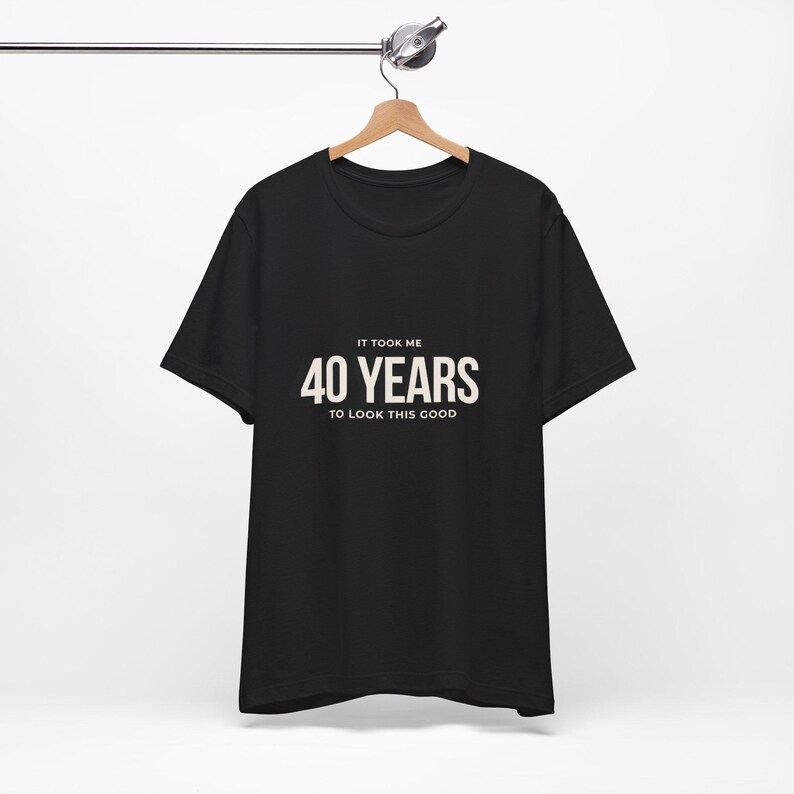 May include: Black t-shirt with the text "IT TOOK ME 40 YEARS TO LOOK THIS GOOD" in white. The shirt is hanging on a wooden hanger attached to a metal bar.