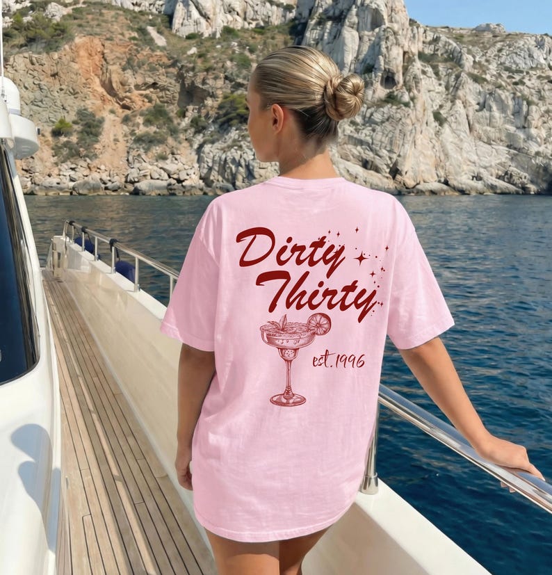 May include: A light pink t-shirt featuring dark red text on the back that reads Dirty Thirty with a graphic of a cocktail glass garnished with a lime slice and the text est. 1996. The design includes small star accents surrounding the text.