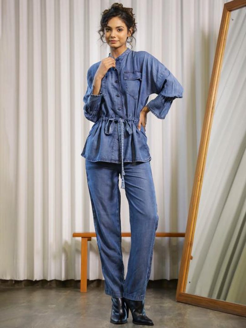 May include: A denim jumpsuit in a light blue wash. The top has a mandarin collar, button-down front, and a drawstring waist. The long sleeves have a flared design. The matching pants are straight-legged. The model is wearing black boots.