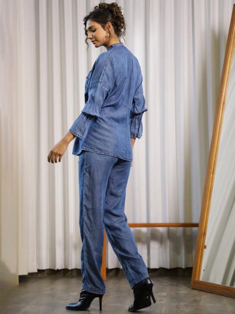 May include: A denim two-piece outfit, consisting of a long-sleeved top and matching pants. The top has ruffled sleeves and a mandarin collar. The pants are straight-legged. The outfit is a medium blue color. The person is wearing black high-heeled boots.