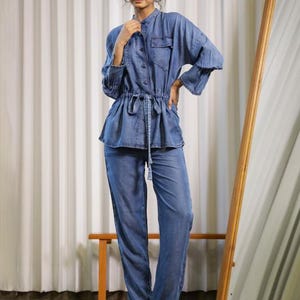May include: A denim jumpsuit in a light blue wash. The top features a mandarin collar, button-down front, and a drawstring waist. The long sleeves have a flared design. The matching pants are wide-legged. The model is wearing black boots.