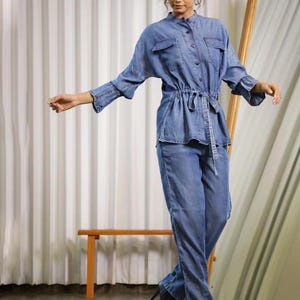 May include: A denim jumpsuit in a light blue wash. The top has a button-down front, two chest pockets, and a cinched waist with a matching belt. The long sleeves have flared cuffs. The pants are straight-legged. The model is wearing black boots.