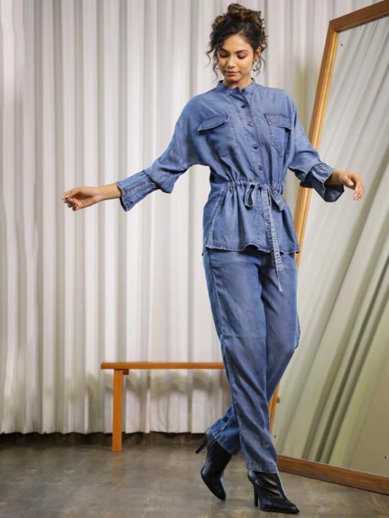May include: A denim jumpsuit in a light blue wash. The top has a button-down front, two chest pockets, and a cinched waist with a matching belt. The long sleeves have flared cuffs. The pants are straight-legged. The model is wearing black boots.