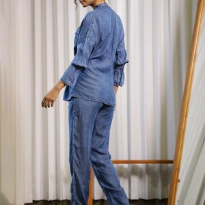 May include: A denim two-piece outfit, featuring a long-sleeved top with ruffled sleeves and matching straight-leg pants. The outfit is a light blue wash. The person is wearing black high-heeled boots. A full-length mirror is in the background.