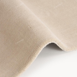 Op de afbeelding: Close-up van een beige tapijt met een zacht, getextureerd oppervlak. Het tapijt is omgevouwen, waardoor de dikte en constructie zichtbaar zijn. De neutrale kleur en de zachte uitstraling suggereren comfort en warmte, geschikt voor de interieurdecoratie.