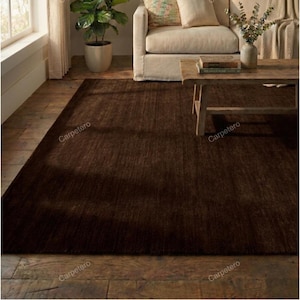 May include: A rectangular, dark brown rug with a textured surface. The rug is placed on a wooden floor in a living room setting, with a sofa and coffee table visible in the background. The rug's color creates a warm and inviting atmosphere.