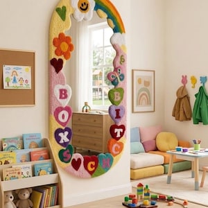 Pode incluir: Um espelho de parede oval com uma moldura espessa, tufada e multicolorida, decorada com várias formas, incluindo um arco-íris, uma flor sorridente, corações e uma borboleta. A moldura exibe as letras B, O, X, S, B, I, T, C, H e C em texto colorido.