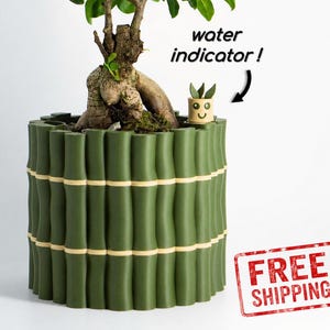 May include: A cylindrical green planter with a textured bamboo-like exterior and light-colored horizontal bands. A small, tan, smiling character figure is placed inside the rim, labeled with the text water indicator! and an arrow.