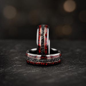 Puede incluir: Dos anillos negros con una incrustación moteada roja y negra y una fina banda dorada. Los anillos se colocan sobre un fondo oscuro, mostrando sus superficies pulidas y reflectantes. Probablemente sean alianzas de boda o accesorios de moda.
