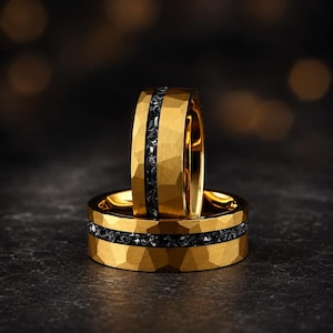 May include: Two gold-coloured rings with a textured surface and a band of black stones. The rings are stacked, with one standing upright. The dark background enhances the rings' details and colour.
