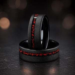 Op de afbeelding: Twee zwarte ringen met een getextureerde afwerking. Elke ring heeft een band van rode, onregelmatig gevormde stenen in een zwart kanaal. De ringen worden gepresenteerd op een donkere ondergrond, waarbij hun contrasterende kleuren en texturen worden getoond.