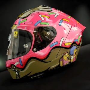 May include: Pink motorcycle helmet with a donut design, featuring sprinkles and a brown frosting base. The helmet has a clear visor and a cartoon illustration of characters. The helmet is a size L.