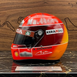 Könnte beinhalten: Ein Miniatur-Rennhelm im Maßstab 1:2 mit einem Farbverlauf in Rot, Orange und Gelb. Er zeigt den Namen M. Schumacher, eine Unterschrift und verschiedene Sponsorenlogos wie Kerakoll, Tic Tac und Shell. Eine Plakette kennzeichnet ihn als 2000 First World Title Ferrari Edition.