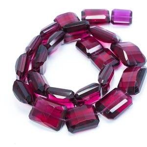 May include: A necklace of faceted, rectangular, deep red gemstones. The gems are arranged in a circular pattern, with a few loose stones visible. The stones have a polished, transparent appearance, reflecting light. The background is white.