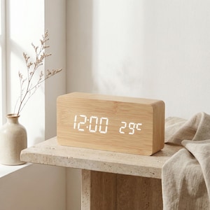 Puede incluir: Un reloj digital rectangular de madera que muestra "12:00" y "29°C" en blanco. El reloj es de madera clara y está sobre una superficie de piedra. Un jarrón con una planta es visible en el fondo.