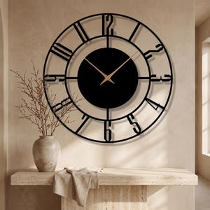 May include: A large, black metal wall clock with Roman numerals and a circular design. The clock features a black center and gold-colored hands. The clock is mounted on a beige wall, with a vase and decorative items in the foreground.