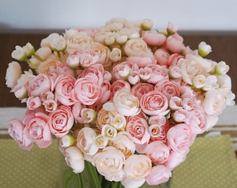 May include: A large bouquet of pink and cream-colored roses in a clear glass vase. The flowers are tightly clustered, with varying sizes of blooms and buds. The vase sits on a light green, polka-dotted surface.
