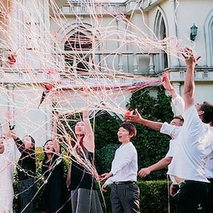 May include: A group of people are gathered outside a building, pulling on streamers that are attached to a balloon. The streamers are pink, white, and blue. The people are smiling and laughing.