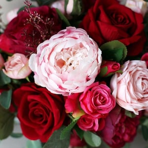 May include: A bouquet of pink, red, and burgundy roses and peonies. The bouquet is arranged in a circular shape and is tied with a ribbon.