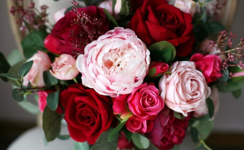 May include: A bouquet of pink, red, and burgundy roses and peonies. The bouquet is arranged in a circular shape and is tied with a ribbon.